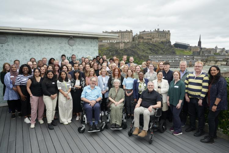 A group photo of many of the people working together to deliver MND-SMART at the 2024 Investigators Gathering in Edinburgh. Picture is taken outside, with a view of Edinburgh castle in the background.  